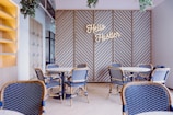 A modern cafe interior with blue and beige chairs surrounding circular tables. A geometric wooden wall feature is present, displaying the neon sign 'Hello Hustler'. Hanging plants and an open shelving unit add decorative elements.