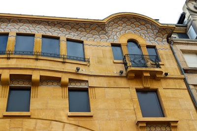 A building facade features warm yellow stonework with large, dark rectangular windows. The upper section is adorned with intricate, colorful mosaic patterns resembling an art deco style. Ornate black metal railings are present, along with decorative moldings and small spotlights affixed to the building.