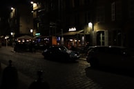 A dimly lit cobblestone street with vintage-style buildings and a small restaurant named 'La Cloche' in view. Street lamps and outdoor seating under umbrellas create a cozy atmosphere. Two cars are parked on the side of the street.