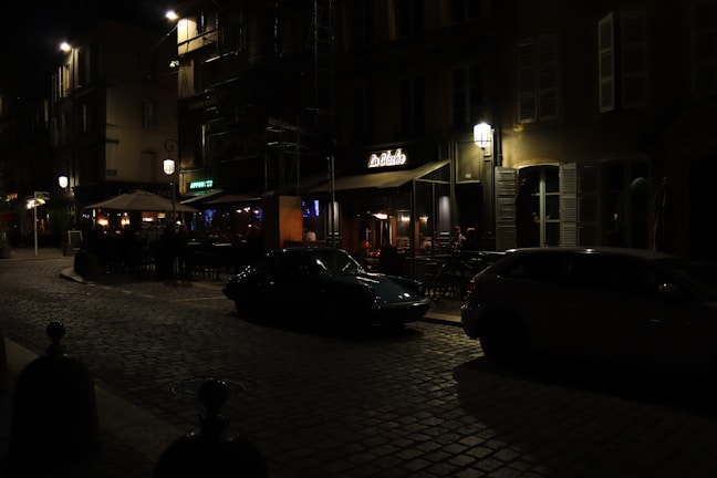 A dimly lit cobblestone street with vintage-style buildings and a small restaurant named 'La Cloche' in view. Street lamps and outdoor seating under umbrellas create a cozy atmosphere. Two cars are parked on the side of the street.