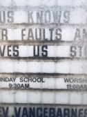 A weathered outdoor sign displays partial religious messages and details about Sunday services. The sign includes the phrases 'KNOWS OUR FAULTS AND STILL LOVES US,' 'SUNDAY SCHOOL 9:30AM,' and 'WORSHIP 11:00AM.' The lettering is somewhat obscured by dirt and wear.