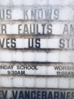 A weathered outdoor sign displays partial religious messages and details about Sunday services. The sign includes the phrases 'KNOWS OUR FAULTS AND STILL LOVES US,' 'SUNDAY SCHOOL 9:30AM,' and 'WORSHIP 11:00AM.' The lettering is somewhat obscured by dirt and wear.