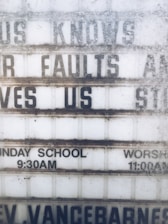 A weathered outdoor sign displays partial religious messages and details about Sunday services. The sign includes the phrases 'KNOWS OUR FAULTS AND STILL LOVES US,' 'SUNDAY SCHOOL 9:30AM,' and 'WORSHIP 11:00AM.' The lettering is somewhat obscured by dirt and wear.