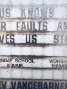 A weathered outdoor sign displays partial religious messages and details about Sunday services. The sign includes the phrases 'KNOWS OUR FAULTS AND STILL LOVES US,' 'SUNDAY SCHOOL 9:30AM,' and 'WORSHIP 11:00AM.' The lettering is somewhat obscured by dirt and wear.