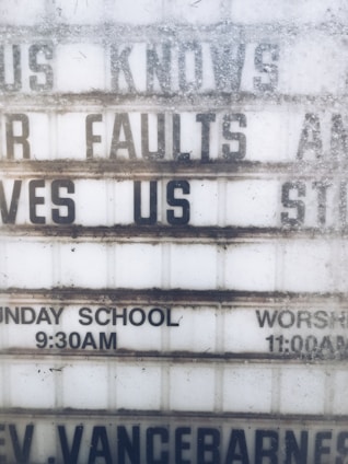 A weathered outdoor sign displays partial religious messages and details about Sunday services. The sign includes the phrases 'KNOWS OUR FAULTS AND STILL LOVES US,' 'SUNDAY SCHOOL 9:30AM,' and 'WORSHIP 11:00AM.' The lettering is somewhat obscured by dirt and wear.