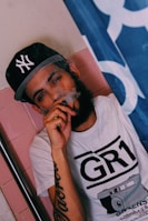 A person wearing a black baseball cap with a logo and a white t-shirt featuring a graphic design of a camera. They are holding a vaping device to their mouth, and smoke is visible around their face. The background includes pink tiles and a partially visible blue and white flag.