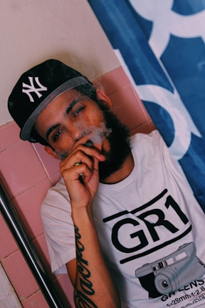 A person wearing a black baseball cap with a logo and a white t-shirt featuring a graphic design of a camera. They are holding a vaping device to their mouth, and smoke is visible around their face. The background includes pink tiles and a partially visible blue and white flag.