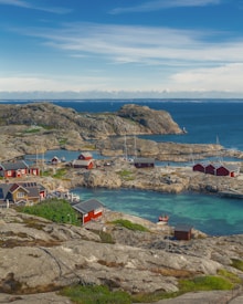 A picturesque coastal scene featuring several red wooden cabins situated on rocky islands surrounded by calm blue waters. In the foreground, rocky terrain and green vegetation can be seen, while the distant horizon shows a clear sky.