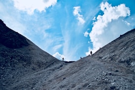 A rugged mountainous landscape with a clear blue sky and scattered clouds above. Several silhouetted figures are walking along a path at the top of a ridge, creating a sense of adventure and exploration.