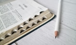 A close-up view of an open book with visible text and black tabs along the side. A white pencil lies next to the book on a light-colored wooden surface.