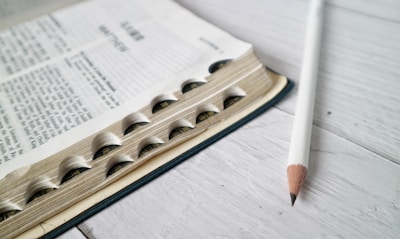 A close-up view of an open book with visible text and black tabs along the side. A white pencil lies next to the book on a light-colored wooden surface.
