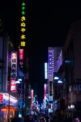 A bustling urban street scene at night, illuminated by a variety of colorful neon signs displaying Japanese characters. The street is crowded with people walking beneath the vibrant lights, conveying a lively nightlife atmosphere. Buildings line the narrow path, each adorned with signage in bright reds, blues, greens, and purples.