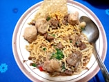 A bowl of noodles mixed with chunks of meat, meatballs, and garnished with chopped green herbs. A crispy cracker is placed at the edge of the bowl, and a metallic spoon rests besides the noodles on the plate. The background is a blue surface with a subtle pattern.