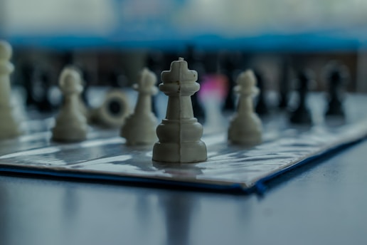 A close-up view of a chessboard featuring white and black chess pieces with a focus on a white king in the foreground, set on a reflective surface. The background is softly blurred, adding depth to the composition.