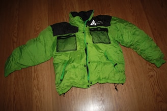 A bright green puffer jacket with black accents is laid flat on a wooden floor. The jacket has two mesh pockets on the front in black, along with other black detailing including the collar and shoulders. It features a printed design on the left chest area.