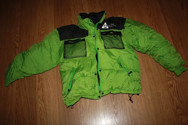 A bright green puffer jacket with black accents is laid flat on a wooden floor. The jacket has two mesh pockets on the front in black, along with other black detailing including the collar and shoulders. It features a printed design on the left chest area.