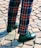A pair of legs wearing brightly colored plaid pants with a pattern of red, blue, and yellow stripes. The pants are paired with dark green leather shoes, and the ground is covered with cobblestone paving.