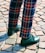 A pair of legs wearing brightly colored plaid pants with a pattern of red, blue, and yellow stripes. The pants are paired with dark green leather shoes, and the ground is covered with cobblestone paving.
