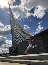 A person is captured mid-air in an energetic leap against a dramatic backdrop of a tall monument with a steep, curved structure that appears to symbolize a rocket. The sky is filled with large, fluffy clouds, and the sunlight enhances the vibrant colors and dynamic composition of the scene.