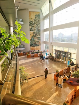 A spacious modern interior features a large vertical painting on a tall white wall. The area includes an open atrium with abundant natural light streaming through large windows on the right. Several people are walking or seated near curved reception desks surrounded by potted plants. The space conveys a sense of openness with plants adding a touch of greenery.