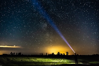 A person stands in a field at night, directing a bright flashlight beam upwards toward the starry sky. The backdrop shows a vast expanse of stars, with the Milky Way visible. The lower horizon features a silhouette of trees against a warm, glowing orange light.