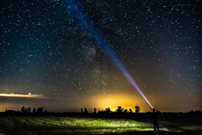 A person stands in a field at night, directing a bright flashlight beam upwards toward the starry sky. The backdrop shows a vast expanse of stars, with the Milky Way visible. The lower horizon features a silhouette of trees against a warm, glowing orange light.