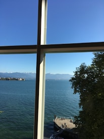 A picturesque lake view framed by window panes, with a blue sky and distant mountains. A tranquil dock is situated at the bottom right, surrounded by lush green foliage.