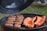 A barbecue grill filled with cooking meats, including sausages and ribs. The grill lid is open, and smoke is gently rising, indicating the food is being cooked. The background shows a hint of green grass, suggestive of an outdoor setting.