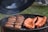A barbecue grill filled with cooking meats, including sausages and ribs. The grill lid is open, and smoke is gently rising, indicating the food is being cooked. The background shows a hint of green grass, suggestive of an outdoor setting.