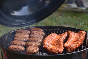 A barbecue grill filled with cooking meats, including sausages and ribs. The grill lid is open, and smoke is gently rising, indicating the food is being cooked. The background shows a hint of green grass, suggestive of an outdoor setting.