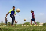 Children are playing soccer in a grassy field. One child in a blue and green jersey is skillfully juggling a soccer ball, while another in a striped jersey focuses on the ball in mid-air. A third child stands nearby, observing. The background features trees, a clear blue sky, and off in the distance, some residential buildings.