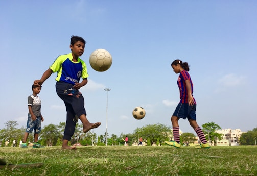 Children are playing soccer in a grassy field. One child in a blue and green jersey is skillfully juggling a soccer ball, while another in a striped jersey focuses on the ball in mid-air. A third child stands nearby, observing. The background features trees, a clear blue sky, and off in the distance, some residential buildings.