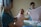 A newborn baby is being held by a caregiver in a medical setting. The baby is wrapped snugly in a pink blanket. Another caregiver in the background is folding a cloth. The atmosphere appears gentle and calm.