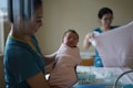 A newborn baby is being held by a caregiver in a medical setting. The baby is wrapped snugly in a pink blanket. Another caregiver in the background is folding a cloth. The atmosphere appears gentle and calm.