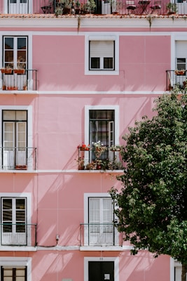 A pink apartment building featuring multiple balconies with potted plants and flowers. The balconies have metal railings, and there's a large tree partially obscuring the bottom right corner. Each floor has large windows with white frames and some are adorned with flowerboxes.