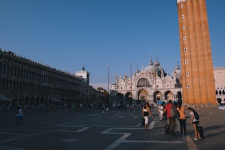 An open plaza with a large, ornate cathedral featuring domes and arches. A tall brick bell tower stands prominently next to the cathedral. Many people are walking around the square, some with luggage, suggesting a tourist area. The sky is clear and blue, indicating a sunny day.