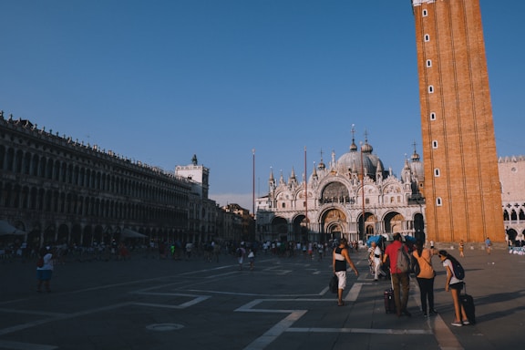 An open plaza with a large, ornate cathedral featuring domes and arches. A tall brick bell tower stands prominently next to the cathedral. Many people are walking around the square, some with luggage, suggesting a tourist area. The sky is clear and blue, indicating a sunny day.
