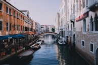 A picturesque canal scene featuring traditional buildings on either side. The buildings exhibit classic Venetian architecture with intricate details and a warm color palette. Small boats are moored along the canal, and a quaint bridge arches over the water in the background. Outdoor seating from a pizzeria lines one side of the canal, enhancing the lively ambiance.
