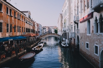 A picturesque canal scene featuring traditional buildings on either side. The buildings exhibit classic Venetian architecture with intricate details and a warm color palette. Small boats are moored along the canal, and a quaint bridge arches over the water in the background. Outdoor seating from a pizzeria lines one side of the canal, enhancing the lively ambiance.