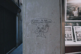 A simple line drawing depicts a face and hand on a textured wall, accompanied by an Italian phrase in a speech bubble. Beside this, there is a window displaying framed photographs, one of which features a couple in a wedding setting.