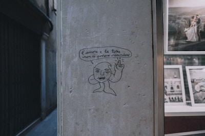 A simple line drawing depicts a face and hand on a textured wall, accompanied by an Italian phrase in a speech bubble. Beside this, there is a window displaying framed photographs, one of which features a couple in a wedding setting.
