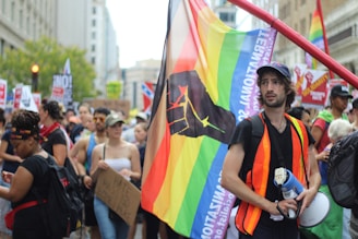 A group of people gather in a street protest, with many holding signs and banners. Prominently, a person wearing a safety vest holds a flag with a rainbow design and a black fist symbol. The mood appears determined and vibrant with diverse participation.
