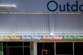 A series of neon lights spelling out 'Doing Things' in various colors is displayed on the exterior of a building. The building facade is a muted blue color with vertical paneling. Part of the word 'Outdoor' is visible at the top. Below the neon lights, there are windows and a partial view of items inside.