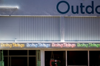 A series of neon lights spelling out 'Doing Things' in various colors is displayed on the exterior of a building. The building facade is a muted blue color with vertical paneling. Part of the word 'Outdoor' is visible at the top. Below the neon lights, there are windows and a partial view of items inside.