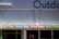 A series of neon lights spelling out 'Doing Things' in various colors is displayed on the exterior of a building. The building facade is a muted blue color with vertical paneling. Part of the word 'Outdoor' is visible at the top. Below the neon lights, there are windows and a partial view of items inside.