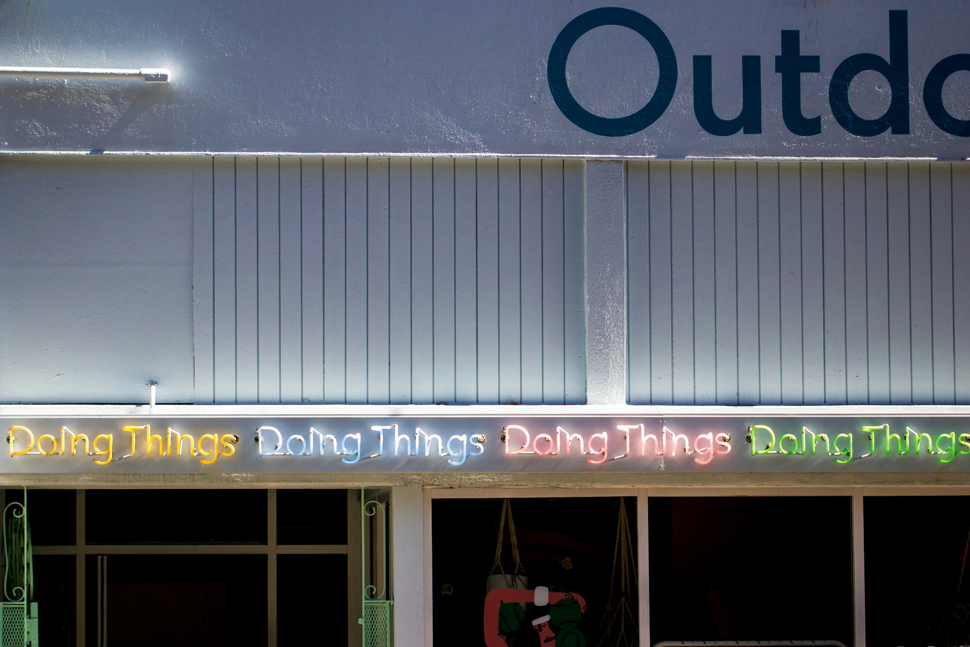 A series of neon lights spelling out 'Doing Things' in various colors is displayed on the exterior of a building. The building facade is a muted blue color with vertical paneling. Part of the word 'Outdoor' is visible at the top. Below the neon lights, there are windows and a partial view of items inside.
