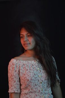 A woman with long, wavy hair is wearing an off-the-shoulder, patterned top with buttons. The background is dark, creating a contrast that highlights her features.