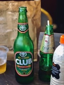 A table display featuring an assortment of beverages. There is a large green glass bottle of Club Premium Lager from Ghana, adorned with branding labels. Next to it is a slightly smaller bottle of Alvaro in a lighter green hue with decorative labeling. Also present is a plastic bottle containing a creamy-looking drink with a label listing ingredients like milk, vanilla, and cinnamon. In the background, a brown paper bag, possibly for takeout, is visible along with a partially visible plastic cup containing a light-colored liquid.