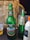 A table display featuring an assortment of beverages. There is a large green glass bottle of Club Premium Lager from Ghana, adorned with branding labels. Next to it is a slightly smaller bottle of Alvaro in a lighter green hue with decorative labeling. Also present is a plastic bottle containing a creamy-looking drink with a label listing ingredients like milk, vanilla, and cinnamon. In the background, a brown paper bag, possibly for takeout, is visible along with a partially visible plastic cup containing a light-colored liquid.
