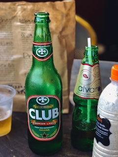 A table display featuring an assortment of beverages. There is a large green glass bottle of Club Premium Lager from Ghana, adorned with branding labels. Next to it is a slightly smaller bottle of Alvaro in a lighter green hue with decorative labeling. Also present is a plastic bottle containing a creamy-looking drink with a label listing ingredients like milk, vanilla, and cinnamon. In the background, a brown paper bag, possibly for takeout, is visible along with a partially visible plastic cup containing a light-colored liquid.